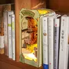 A wooden book nook model with a "Falling Sakura" theme, illuminated from within, placed on a bookshelf between books. Náhled obrázku produktu
