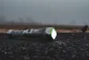 A flashlight lies on a dark, textured surface, possibly asphalt, with a blurred, overcast background suggesting an outdoor environment. Produktbild-Vorschau