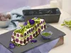 A terrine-style dish made with purple potatoes and peas, garnished with asparagus and edible flowers, served on a slate platter with a small bowl of green sauce. Náhled obrázku produktu