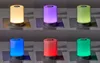 A collage of six images showing a cylindrical LED table lamp in different colors: yellow, purple, light green, blue, red, and dark green. Each lamp is placed on a square platform. Produktbild-Vorschau
