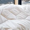A close-up view of a white comforter with a diamond-stitched pattern, set against a blurred background of a snowy mountain village visible through a window. Produktbild-Vorschau