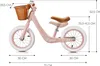 A pink balance bike with a wicker basket and brown seat, shown with dimensions indicated by lines and text. Produktbild-Vorschau