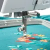 A close-up view of a sewing machine needle and embroidery hoop with floral embroidery. Produktbild-Vorschau
