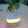 A modern white and gray oval shaped device with a white glowing light strip around its middle sits on a light wood grain table. Produktbild-Vorschau