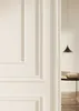A close-up view of a white wall with decorative molding, with a glimpse into another room with a hanging lamp and a table with books. Anteprima dell’immagine prodotto