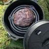 A piece of meat is being cooked in a cast iron pot with a rack inside, set on a mossy ground. Produktbild-Vorschau
