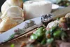 A close-up of a Laguiole knife with a decorative handle, placed on a wooden board with garlic and a portion of food. Voir les images du produit