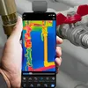 A hand holds a smartphone displaying a thermal image of pipes, with a thermal camera attachment on top of the phone. Plumbing fixtures are visible in the background. Voir les images du produit