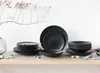 A set of black stoneware plates and bowls are arranged on a wooden shelf, with a decorative branch and stones in the foreground. Produktbild-Vorschau