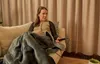 A woman sits on a couch wrapped in a blanket, holding a remote control. Podgląd zdjęcia produktu
