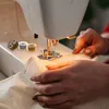 A close-up shot of a person's hands using a sewing machine to stitch fabric. Several spools of thread are visible in the background. Produktbild-Vorschau