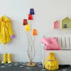 A colorful floor lamp with multiple shades stands in a child's room next to a yellow raincoat and boots, a white bench with a pink cushion, and a yellow drawstring bag. Produktbild-Vorschau