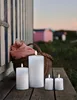 Four white LED candles of varying sizes are placed on a wooden surface in front of a beach hut and a basket with a pink cloth. Produktbild-Vorschau