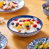A close-up shot of a fruit salad in a blue and white patterned bowl, with other plates of food and dishes visible in the background. Produktbild-Vorschau