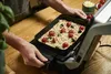 A person is preparing a rectangular pizza in a black baking tray on a wooden table. Produktbild-Vorschau