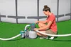 A woman is kneeling next to a pool sand filter system, adjusting a valve. The pool is in the background and the system is on a grassy area. Produktbild-Vorschau