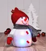 A decorative LED snowman figurine with a red hat and scarf, set against a white, slightly textured background with a silhouette of a Christmas tree. Red Christmas ornaments are visible on the left and right sides. Produktbild-Vorschau