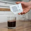 A hand is pouring yeast and nutrient mixture from a white packet into a glass of dark liquid. Produktbild-Vorschau