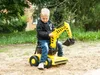 A young boy is playing with a yellow ride-on toy excavator in a sandy area with trees and a building in the background. Podgląd zdjęcia produktu