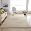 A beige rug is placed on a wooden floor in a living room setting, with a sofa, chair, and plant visible. Podgląd zdjęcia produktu