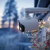 A white security camera covered in snow, decorated with colorful Christmas lights, in a winter outdoor setting. Produktbild-Vorschau
