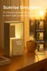 A sunrise simulation alarm clock is shown on a wooden table next to a window with the sun setting. Produktbild-Vorschau