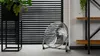 A floor fan is placed on a carpeted floor in front of black horizontal blinds. To the left is a black planter with a green plant. To the right is a metal shelf unit with wooden shelves. Voir les images du produit