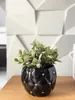 A black ceramic pot with decorative rhinestones and a green plant sits on a wooden surface in front of a textured white wall. Produktbild-Vorschau