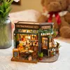 A miniature DIY restaurant model with a teddy bear sitting at a table, placed on a table with a vase of flowers and a large teddy bear in the background. Voir les images du produit