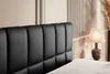 A close-up view of a black upholstered headboard of a bed, with a bedside lamp and decorative dried grass in the background. Produktbild-Vorschau