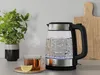 A glass electric kettle boiling water sits on a kitchen counter next to a cup of tea and a plant. Podgląd zdjęcia produktu