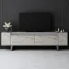 A modern TV stand with a television set on top, placed against a dark wall with decorative paneling. The TV stand has a stone-like finish and metal legs. There are decorative items on the stand, including a vase of flowers and candles. Voir les images du produit