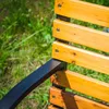 Close-up of a wooden park bench with black metal accents, set against a blurred green grassy background. Anteprima dell’immagine prodotto