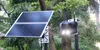 A solar-powered camera with a solar panel mounted on a pole next to a tree. Podgląd zdjęcia produktu
