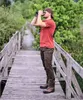 A man is standing on a wooden bridge in a natural environment, looking through binoculars. Produktbild-Vorschau