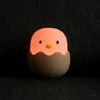 A cute chick-shaped night light is sitting in a gray eggshell on a dark gray textured surface. Produktbild-Vorschau