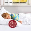 A young boy sleeps peacefully in bed next to a red analog alarm clock. Produktbild-Vorschau