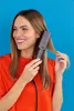 A woman is using a hair straightening brush on her hair against a blue background. Produktbild-Vorschau