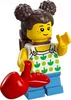 A LEGO minifigure of a girl with brown hair styled in two buns, wearing a white top with green leaf patterns and blue overalls, holding a red object that resembles lips. Podgląd zdjęcia produktu