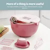 A pink multi-functional kitchen grater and bowl set with various attachments is displayed on a white countertop. Produktbild-Vorschau