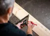 A person is using a black triangular ruler to mark a line on a wooden plank. Voir les images du produit
