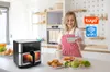 A woman in a kitchen is using a smartphone to control a smart air fryer. There are also some food items on the counter. Produktbild-Vorschau