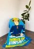 A blue fleece blanket with a cartoon character of Fireman Sam printed on it, draped over a round object in a room with a plant in the background. Produktbild-Vorschau