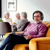 A man wearing headphones and holding a remote control sits on a yellow couch, while three women are in the background at a table. Produktbild-Vorschau