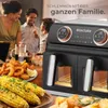 A family is gathered around a table with food, and a dual-chamber air fryer is prominently displayed in the foreground. Produktbild-Vorschau