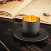 A black espresso cup with a gold interior sits on a black saucer next to an open book. Náhled obrázku produktu