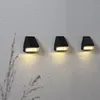 Three black solar wall lights mounted on a textured gray wall, with a blurred plant in the foreground. Produktbild-Vorschau