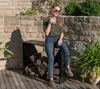 A woman is sitting on a firewood rack next to a stone wall, holding a drink. Produktbild-Vorschau