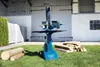 A blue log splitter stands on a grassy lawn in front of a large building with white walls and large double doors. Several logs are stacked to the right of the splitter, and two logs are placed to the left. Produktbild-Vorschau
