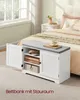 A white storage bench with a gray cushion is placed in a bedroom next to a bed. The bench has two doors, one open, revealing shelves with folded items. A teacup sits on a book on the bench's surface. Produktbild-Vorschau